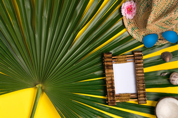 Summer composition with photo frame, green palm leaves, hat and sunglasses on a yellow background. Artwork mockup with copy space