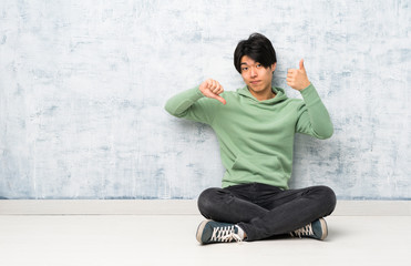 Asian man sitting on the floor making good-bad sign. Undecided between yes or not