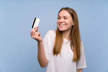 Young woman over blue wall holding a credit card and thinking