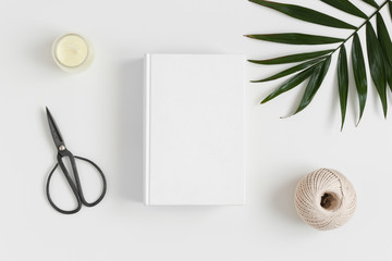 Top view of a white book mockup with  workspace accessories and a palm leaf on a white table.