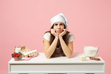 Sad tired female chef cook confectioner or baker in apron white t-shirt, toque chefs hat cooking cake or cupcake at table isolated on pink pastel background in studio. Mock up copy space food concept.