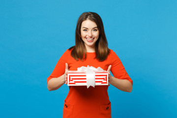 Beautiful young woman wearing red orange dress hold red striped present box with gift ribbon isolated over trendy blue background studio portrait. People lifestyle fashion concept. Mock up copy space.