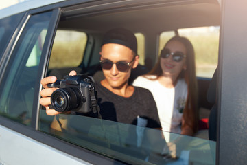 Blogger in glasses filming his travel vlog on professional camera on road trip. Man photographer taking photos out of open window with girlfriend nearby. Multiracial hipsters couple on rear car seat.