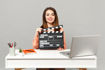 Young beautiful woman holding classic black film making clapperboard sit and work at desk with pc laptop isolated on gray background. Achievement business career lifestyle concept. Mock up copy space.