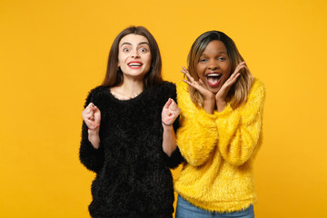 Two young women friends european and african american in black yellow clothes standing posing isolated on bright orange wall background, studio portrait. People lifestyle concept. Mock up copy space.