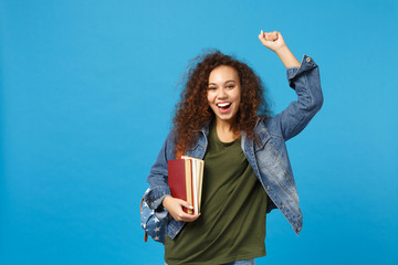 Young african american girl teen student in denim clothes, backpack hold books isolated on blue wall background studio portrait. Education in high school university college concept. Mock up copy space