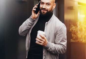 Portrait of smiling man talking on cell phone and drinking coffee on city street, bearded hipster guy calling friends. Lifestyle.