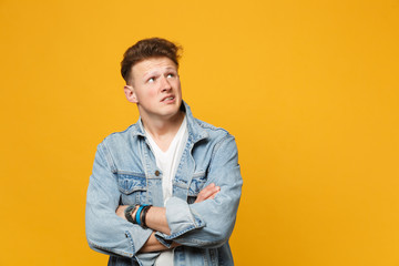 Portrait of disturbed young man in denim casual clothes looking up aside, holding hands crossed isolated on yellow orange background. People sincere emotions lifestyle concept. Mock up copy space.