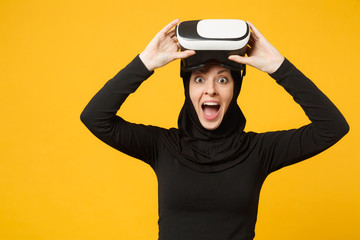 Young arabian muslim woman in hijab black clothes watching in headset of vr virtual reality isolated on yellow wall background, studio portrait. People religious lifestyle concept. Mock up copy space.