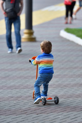 Fototapeta premium Boy child in blue jeans and sneakers riding a scooter. back view