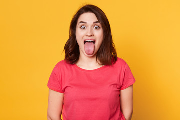 Portrait of beautiful young female with dark hair, has pleasant appearance, shows tongue, having fun with friends or family, wearing red casual t shirt. Beauty, entertainment and youth concept.
