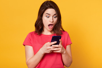 Naklejka premium Studio shot of stupefied Caucasian brunette woman posing with widely open mouth, reads incredible news in Internet or shocking messagw from friwnd, holds mobile phone, dressed red casual tshirt.
