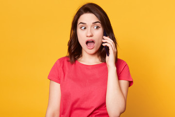 Close up portrait of amazed beautiful girl in casual style, has dark hair, standing and speaking on mobile smart phone and looking with shocked face and open mouth isolated over yellow background.