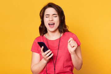 Adorable emotional beautiful young lady standing isolated over yellow background in studio, closing her eyes, holding smartphone, being in big earphones, listening to favorite music, dancing.