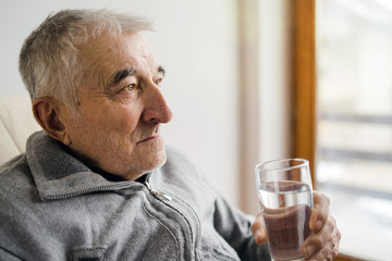 Senior man sitting holding glass of water looking sad by the window at the retirement nursing home