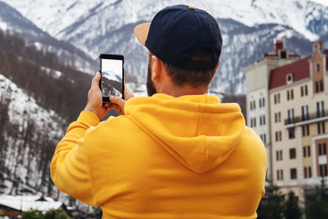 Back view. Bearded male tourist in yellow hoodie and cap stands on background of high snowy mountains, makes photos on smartphone.