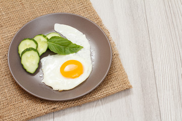 Plate with fried egg, fresh cut cucumber and leaf of basil on sackcloth.