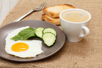 Plate with fried egg, fresh cut cucumber and leaf of basil, fork, cup of black coffee and toasts.