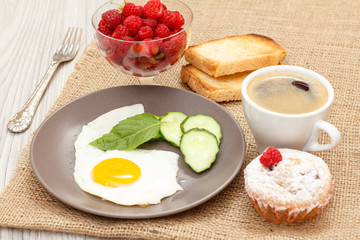 Plate with fried egg, fresh cut cucumber and leaf of basil, muffin, cup of black coffee, fork, toasts and glass bowl with fresh raspberries