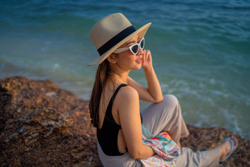 relaxing time concept with traveler girl posing smile in the sea beach in beautiful sunset time by wear sunglass and hat by sit on the stone