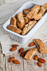 Cookies cantuccini in white ceramic bowl and almond seeds on linen napkin closeup on ructic wooden table background.