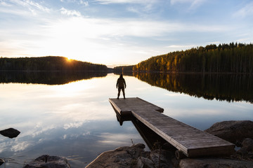 Fototapeta na wymiar Lonely man standing on the edge of the wooden pier looking at the calm lake and colorful forest on the other side