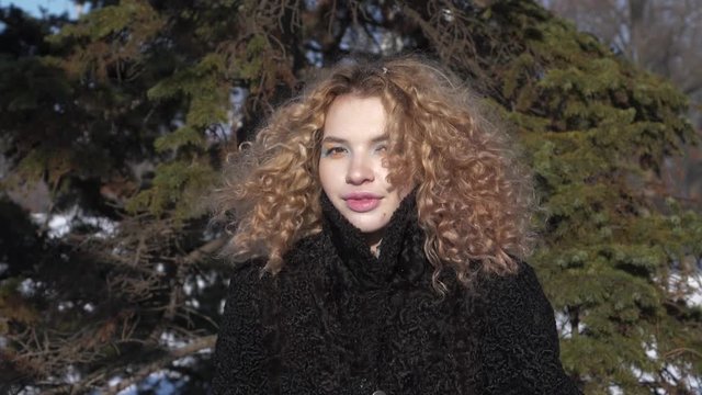 A young woman with a curly haircut in a black coat throws snow over herself Sunny winter day