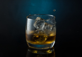 A glass of whiskey with ice on a dark color photo with splashes from falling ice