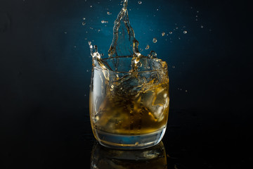 A glass of whiskey with ice on a dark color photo with splashes from falling ice