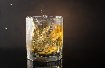A glass of whiskey with ice on a dark color photo with splashes from falling ice