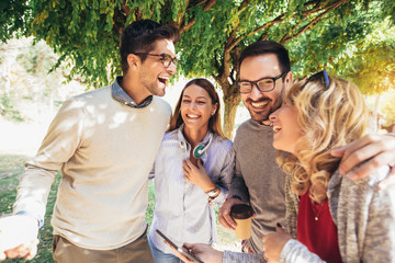 Four happy smiling young friends walking outdoors in the park holding digital tablet