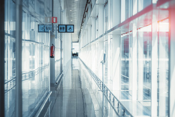A long modern glass and chrome corridor stretching into vanishing point indoors of an airport terminal or a contemporary railway station depot, with a restroom and fire extinguisher on the left side