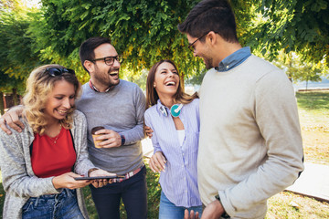 Four happy smiling young friends walking outdoors in the park holding digital tablet
