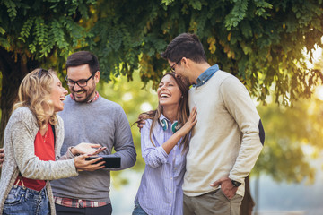 Four happy smiling young friends walking outdoors in the park holding digital tablet