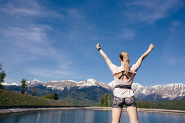 Fototapeta na wymiar Rear view of beautiful fit woman athlete rejoices lifted arms upwards and looking at beautiful landscape with blue sky mountains and lake on a warm summer day. Concept of achieving goals and winner