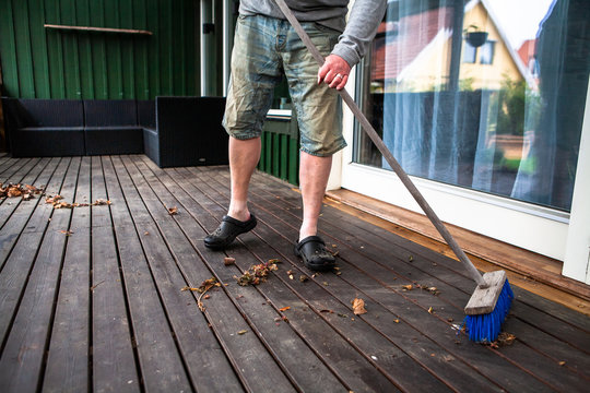 A man wearing jean shorts is sweeping a wooden porch from dry autumn leaves and dirt with a broom. The reflection of a house can be seen in the window. Spring cleaning.