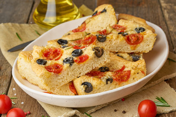Focaccia, pizza in plate with tomatoes, olives and rosemary. Chopped Italian flat bread. Side view, wooden table