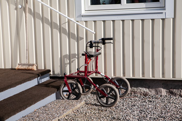 A walker with wheels is parked on the gravel outside a home. The wheels are big for use on uneven terrain outdoors. The walker has brakes, a seat and a shelf for transporting personal belongings.