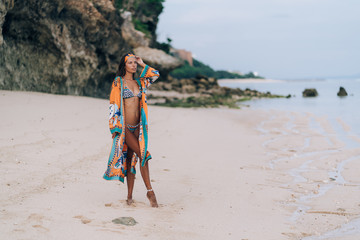 Beautiful sexy girl in swimwear walking on sandy beach with rock on background