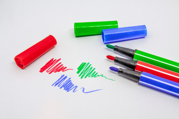 A part of green, blue and red open felt-tip pens and covers from them are near a number of lines drawn by them on a white background.