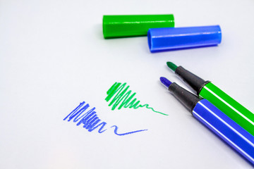 A part of green and blue open felt-tip pens and covers from them are near a number of lines drawn by them on a white background.