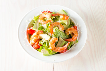 Fresh shrimp salad made of tomato, ruccola, avocado, prawns, chicken breast, arugula, crackers and spices. Caesar salad in a white, transparent bowl on wooden background