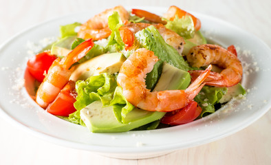 Fresh shrimp salad made of tomato, ruccola, avocado, prawns, chicken breast, arugula, crackers and spices. Caesar salad in a white, transparent bowl on wooden background