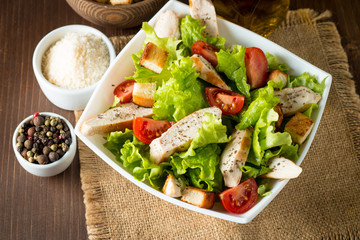 Fresh salad made of tomato, ruccola, chicken breast, eggs, arugula, crackers and spices. Caesar salad in a white, transparent bowl on wooden background