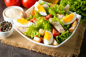 Fresh salad made of tomato, ruccola, chicken breast, eggs, arugula, crackers and spices. Caesar salad in a white, transparent bowl on wooden background