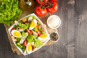 Fresh salad made of tomato, ruccola, chicken breast, eggs, arugula, crackers and spices. Caesar salad in a white, transparent bowl on wooden background