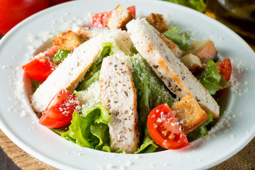 Fresh salad made of tomato, ruccola, chicken breast, eggs, arugula, crackers and spices. Caesar salad in a white, transparent bowl on wooden background