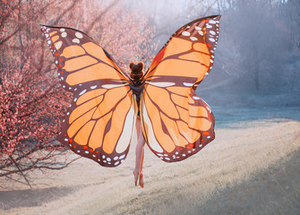 A monarch butterfly enjoys flying. A young woman with a perfect body, long legs flies up into the sky. Huge, painted wings opened like a moth. Art photo from the back, without a face. Sun rays