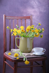 A bouquet of dandelions in a vase and a cup of tea on a chair.