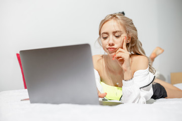 Young girl blogger dressed in fashionable clothes lying on the bed next to the camera and working...